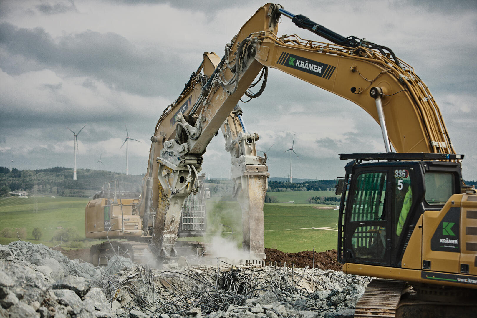 Zwei große gelbe Bagger entfernen Beton und Bewehrungsstahl auf einer Baustelle. Staub wirbelt aus den Trümmern auf. Im Hintergrund sind Windräder und grüne Felder unter einem bewölkten Himmel zu sehen.