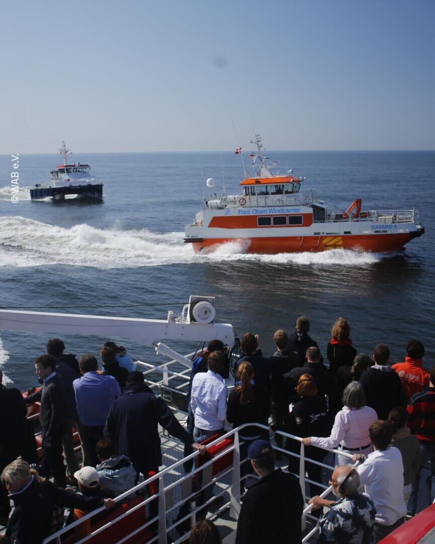 Menschen auf einer Fähre beobachten zwei Arbeitsboote auf ruhiger See unter klarem Himmel. Die Boote hinterlassen beim Vorbeifahren Kielwasser, und die Szene lässt auf eine Vorführung oder eine Veranstaltung auf See schließen.