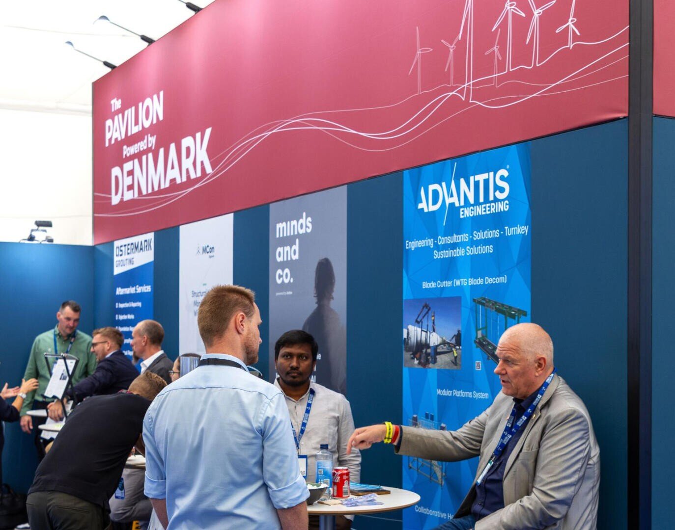 Eine Gruppe von Personen unterhält sich an einem Messestand unter einem roten Schild mit der Aufschrift The Pavilion Powered by Denmark. Im Hintergrund sind Werbebanner und Informationsmaterial zu sehen.