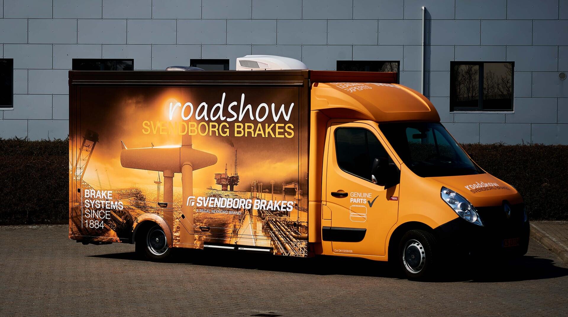 Ein gelber Roadshow-Van, der vor einem Gebäude geparkt ist und auf dessen Seiten in großen Lettern für Svendborg Brakes geworben wird und auf dem Bilder von Industrieszenen aufgedruckt sind.