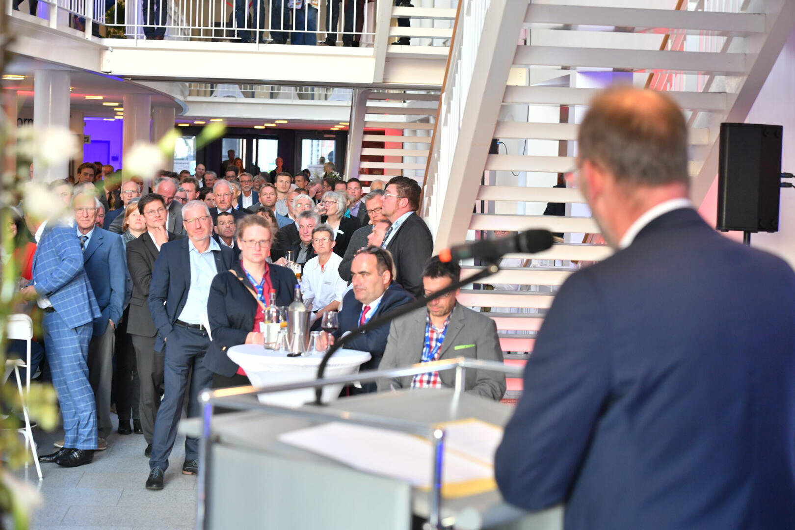 Ein Mann im Anzug spricht an einem Podium zu einem großen, aufmerksamen Publikum in einem modernen, hell erleuchteten Raum mit weißen Treppen und Balkongeländern. Die meisten Anwesenden stehen und tragen Business-Kleidung.