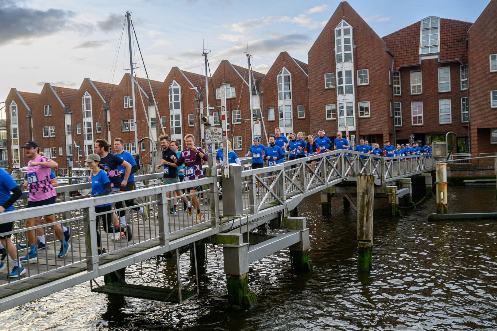 A large group of runners in colorful sportswear cross a narrow bridge over the water, with brick buildings with high windows in the background. The sky is partly cloudy.