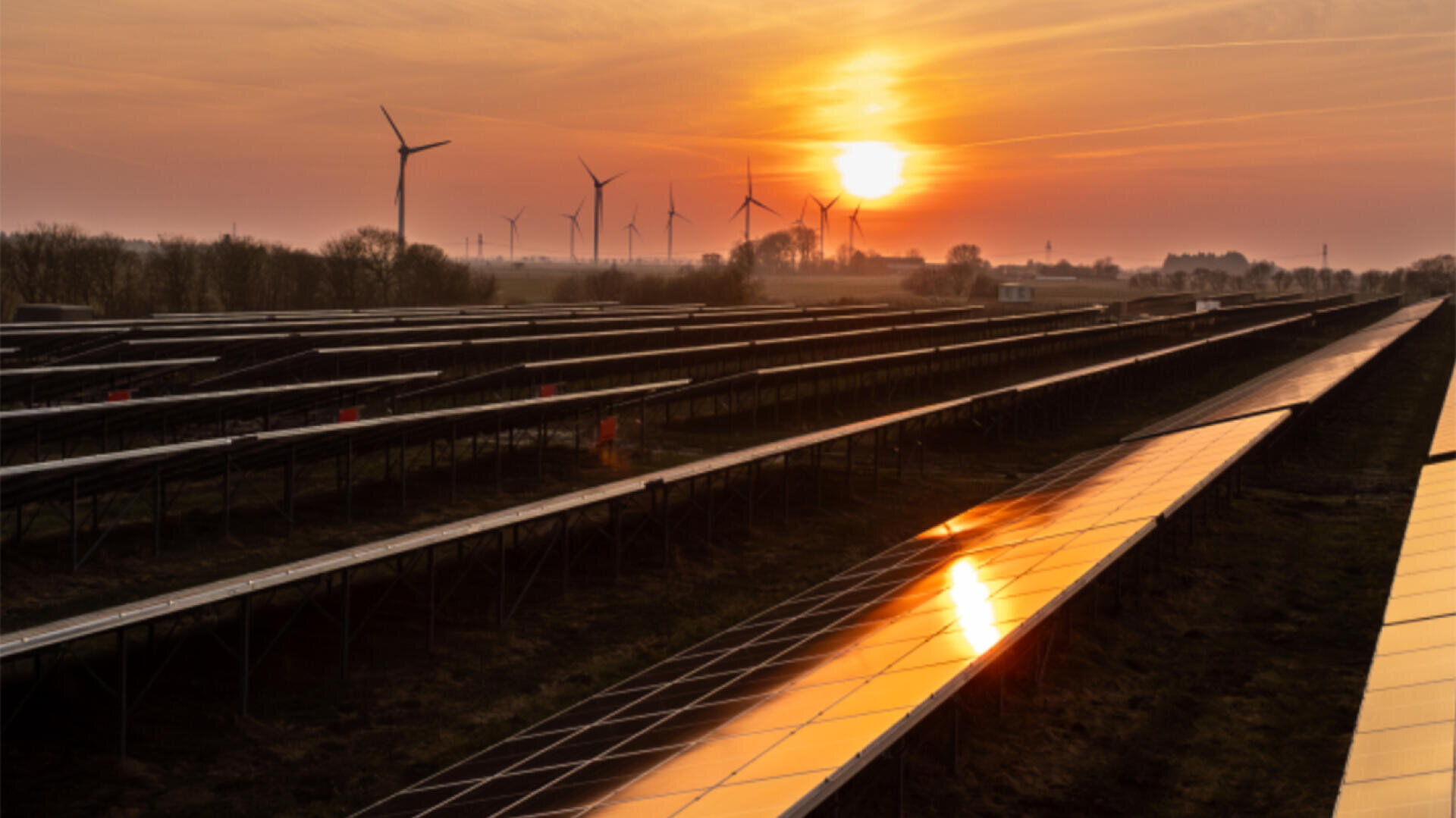 Rows of solar modules and wind turbines in a field at sunset, with the sun reflecting on the modules and the sky in the background turning orange.
