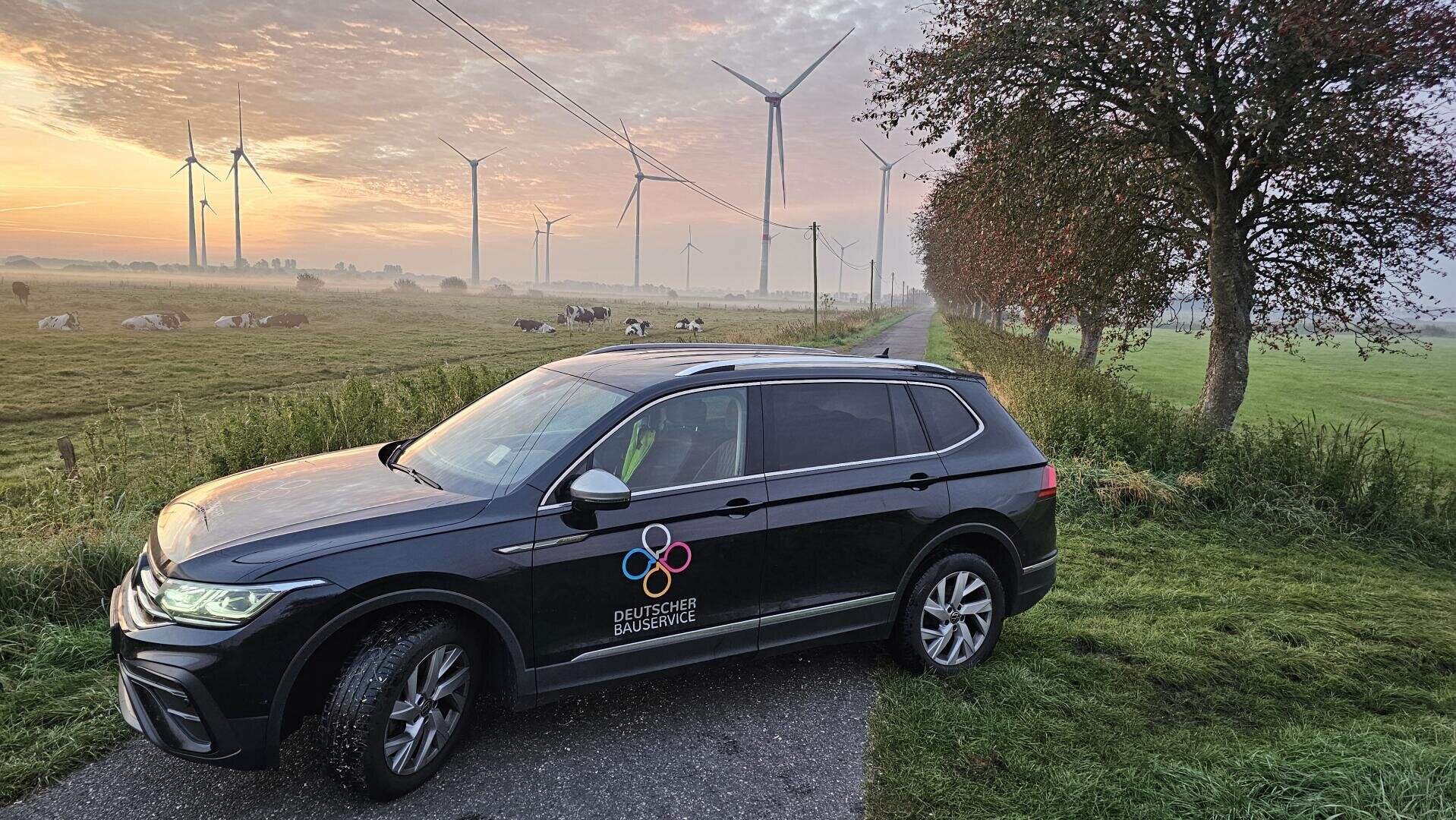 A black SUV bearing the German Energy Service logo is parked on the grassy side of the road near a cow pasture. Wind turbines and power lines can be seen in the background, under a morning sky with orange clouds.