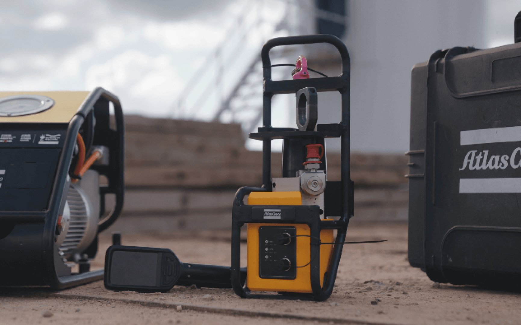 Ein Industriegerät mit gelb-schwarzem Gehäuse steht auf sandigem Boden, umgeben von anderen Maschinen und einem Hartschalenkoffer mit der Aufschrift „Atlas Copco“. Im Hintergrund sind Holzbretter und eine verschwommene Struktur zu sehen.