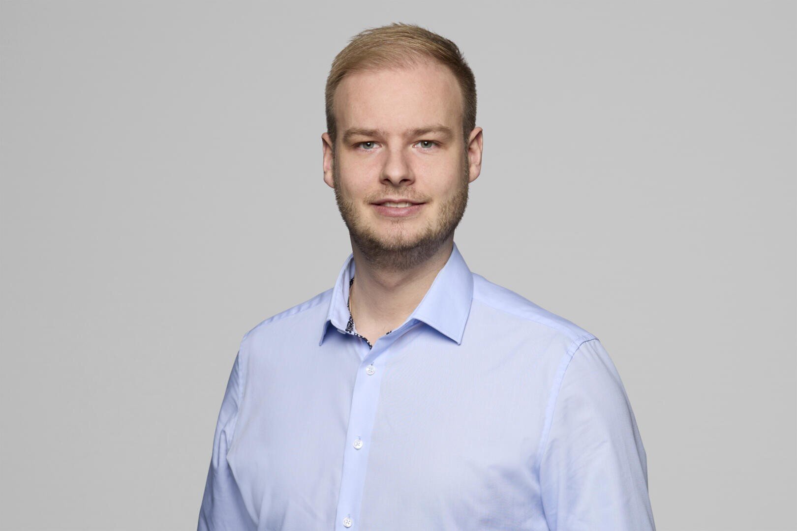 A man with short blonde hair and a trimmed beard is wearing a light blue button-up shirt. He is smiling slightly and posing against a plain, light gray background.