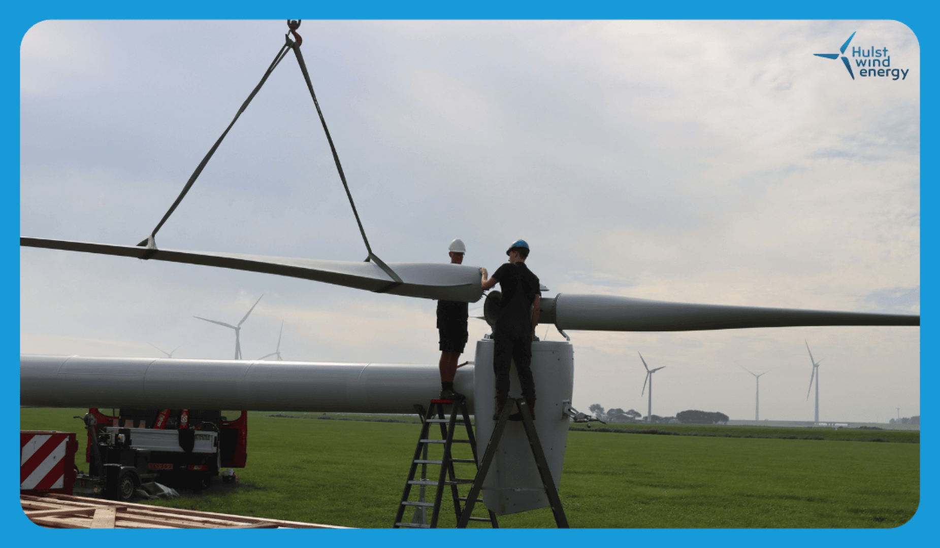 Two workers in protective clothing stand on ladders and use a crane to assemble a wind turbine blade on a meadow with several wind turbines in the background. The sky is partly cloudy. The Hulst Wind Energy logo is in the top right corner.