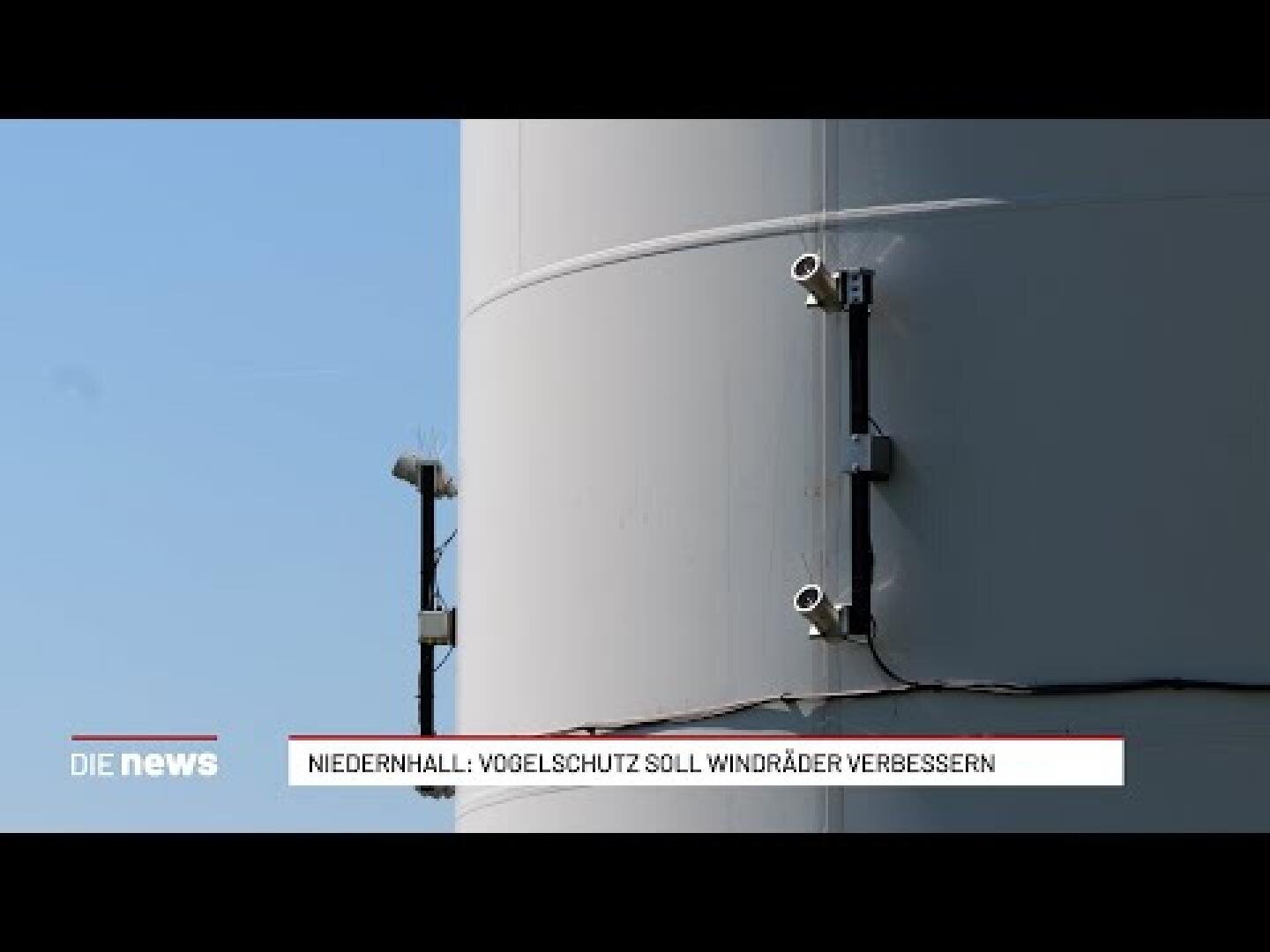 Nahaufnahme von Kameras, die an der Seite eines Windradturms angebracht sind, mit einem klaren blauen Himmel im Hintergrund. Ein Nachrichtenticker am unteren Bildrand berichtet über Vogelschutz und Windkraftanlagen in Niedernhall.