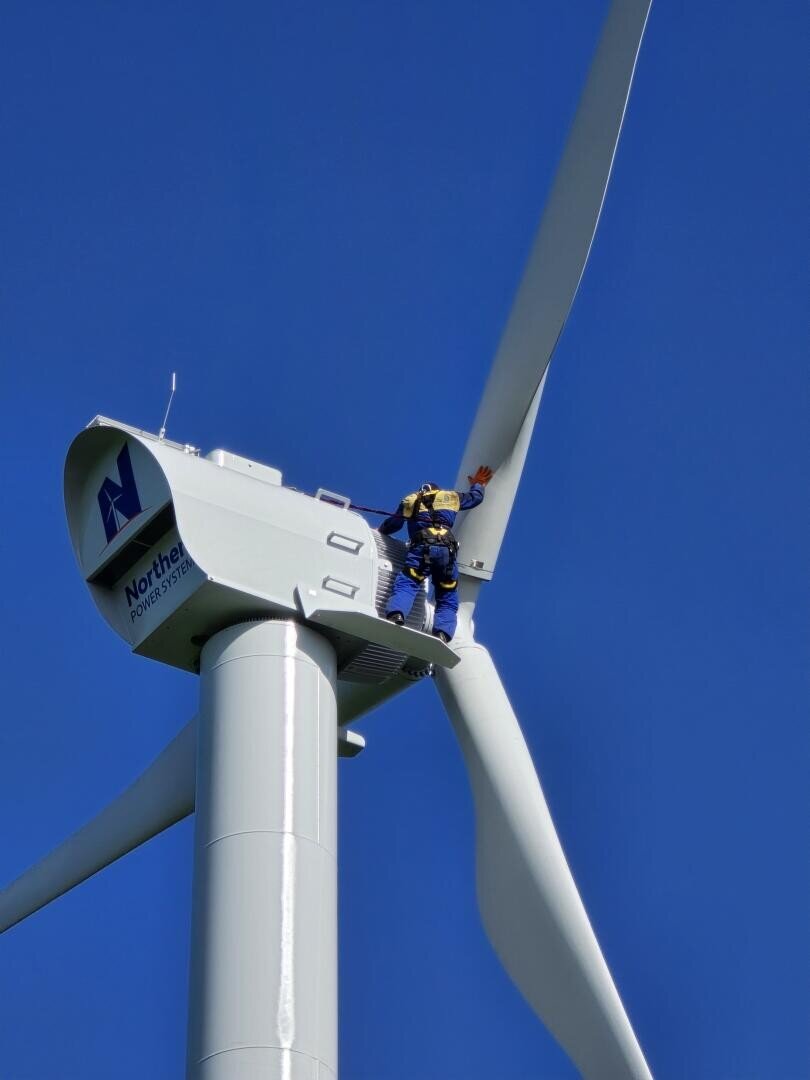 A worker in safety gear stands on top of a wind turbine, reaching out to touch one of the blades against a clear blue sky.