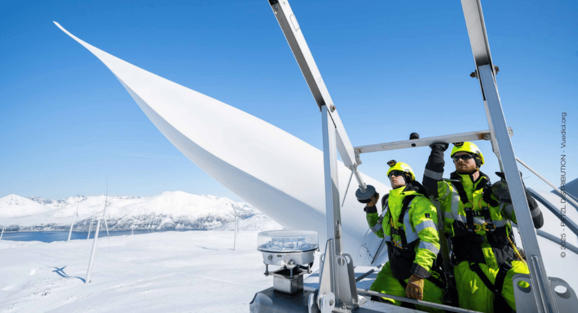 Two workers in bright safety gear inspect a wind turbine in a snowy landscape, with mountains and other wind turbines visible under a clear blue sky.