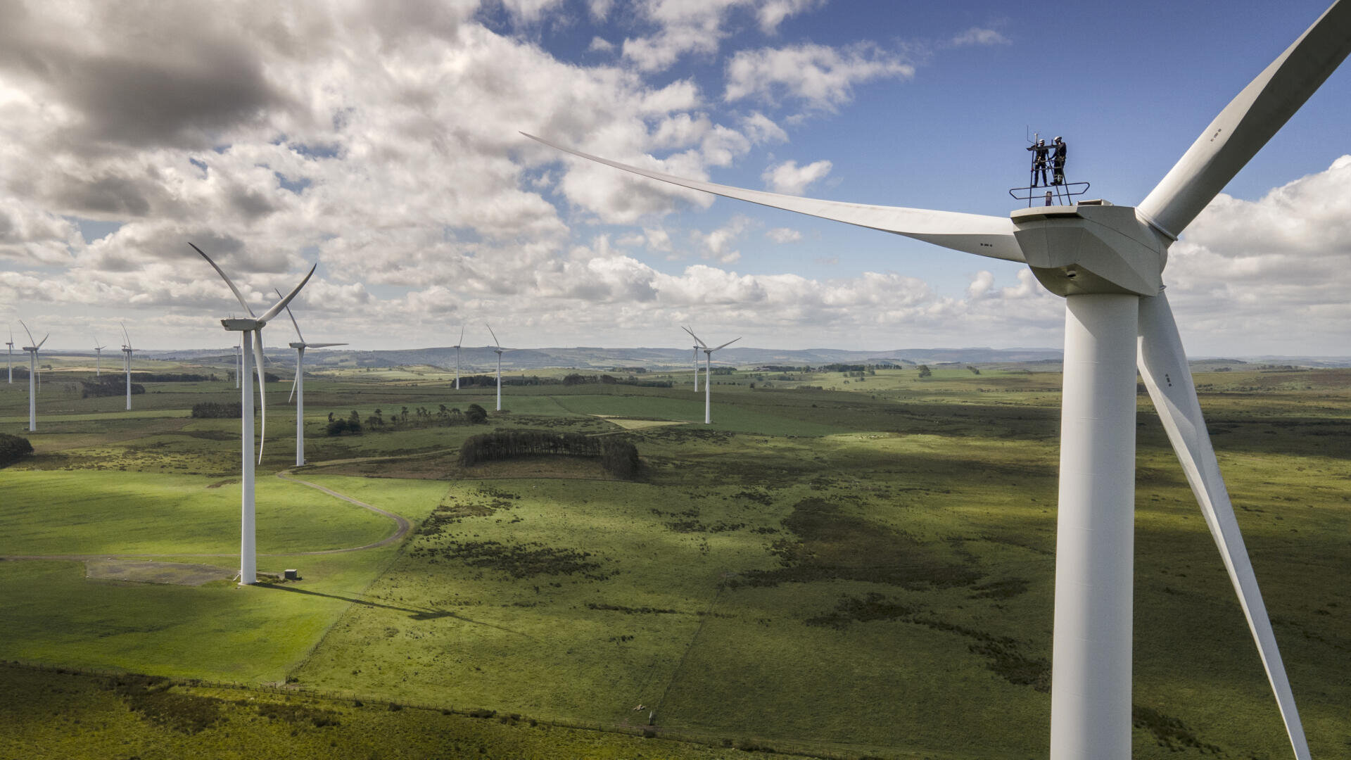 Weite Sicht auf einen Windpark mit mehreren großen weißen Windturbinen auf grünen Feldern unter einem teilweise bewölkten Himmel. Zwei Personen stehen auf der nächstgelegenen Turbine, um sie zu warten.
