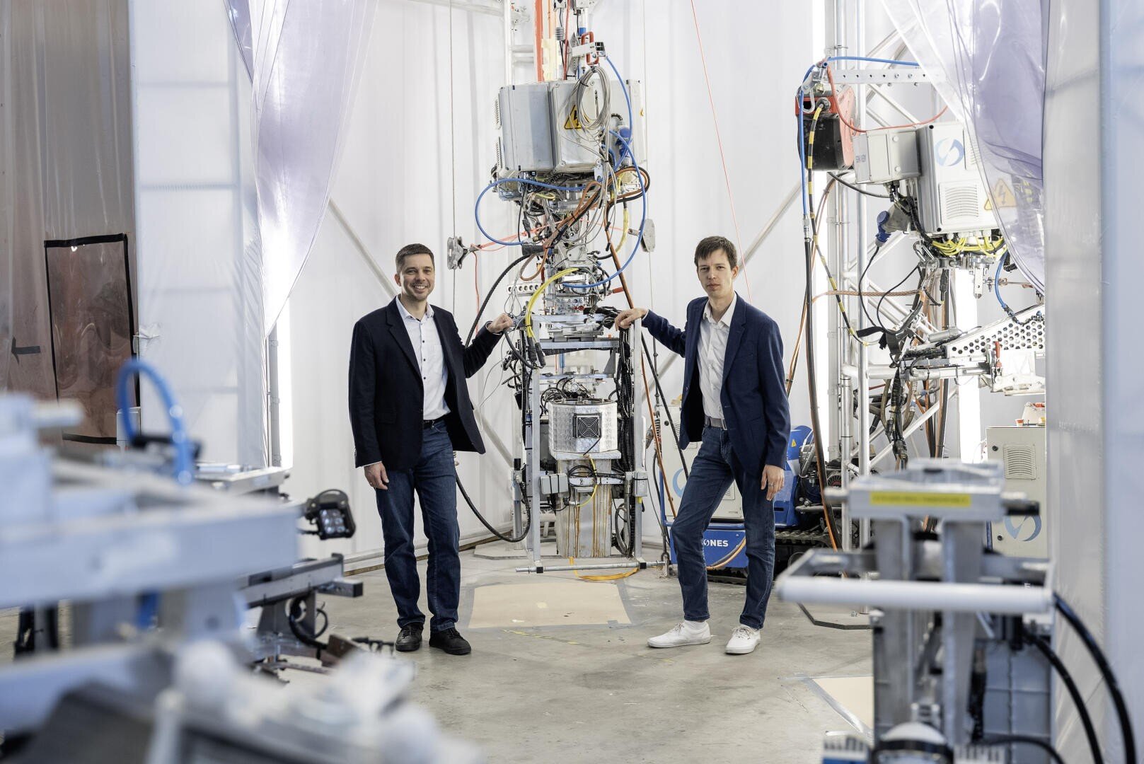 Two men in suits stand in a bright laboratory, each holding part of a tall, complex machine with wires and components. Various technical equipment and cables surround them in the workspace.