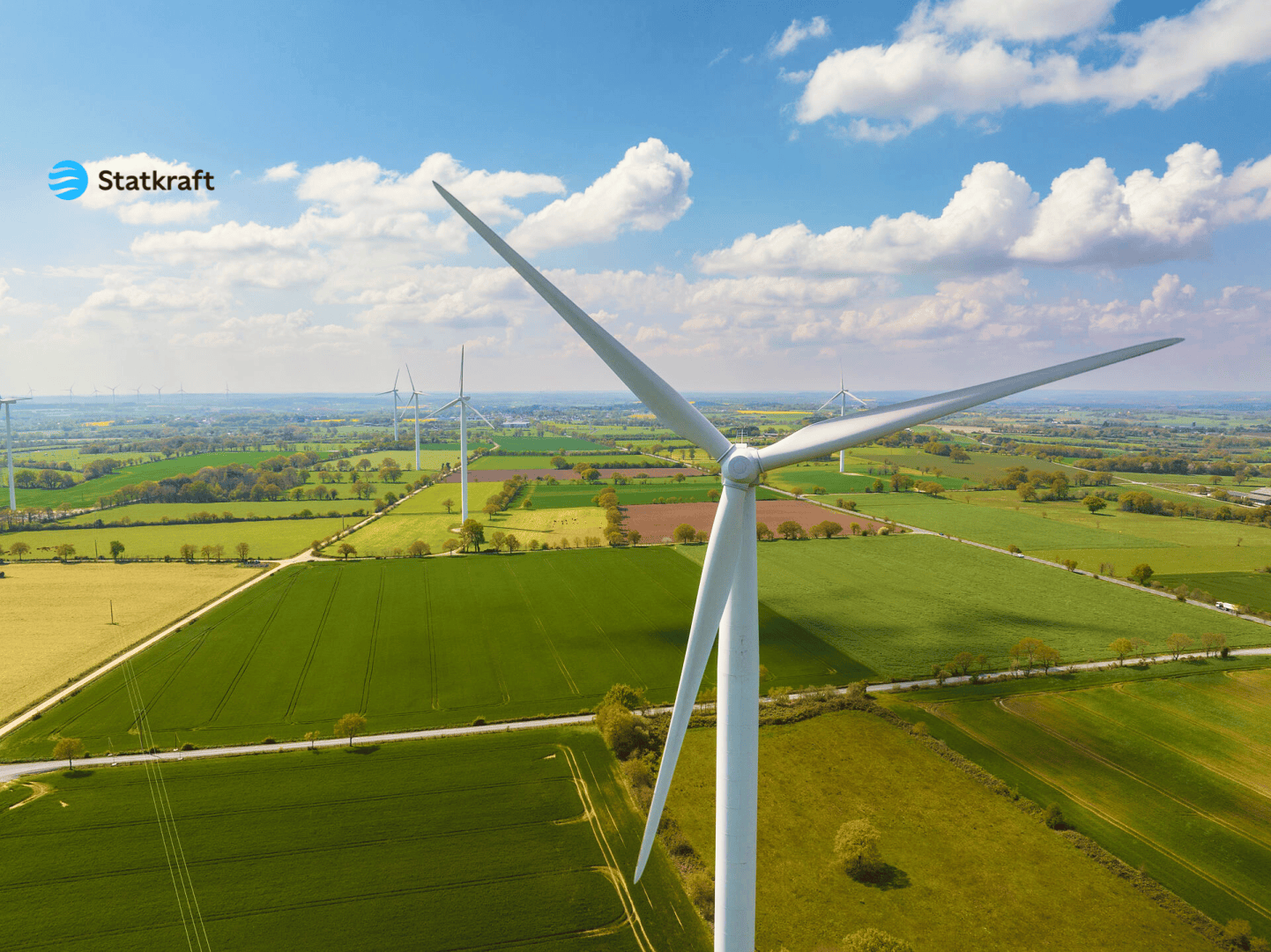 A large wind turbine stands in a green, open landscape with fields and roads under a partly cloudy sky. Several other wind turbines can be seen in the distance. The Statkraft logo appears in the top left corner.