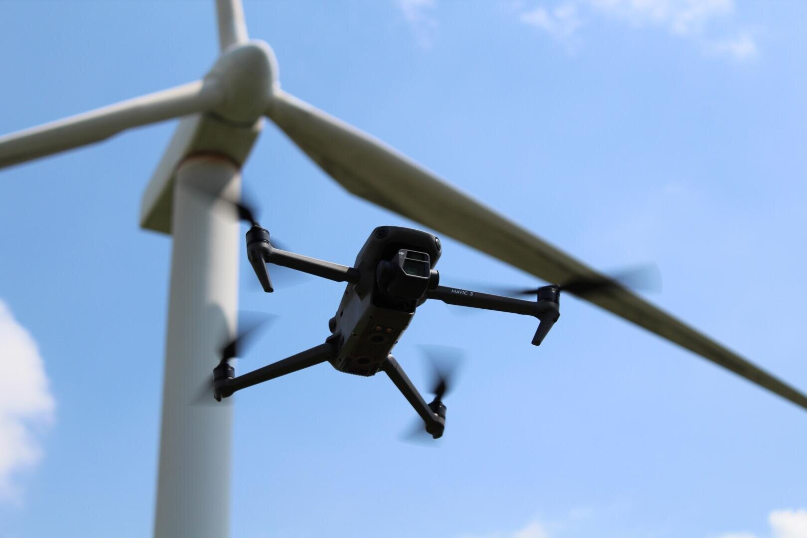 A black drone with spinning propellers flies near the blade of a large white wind turbine against a bright blue sky.