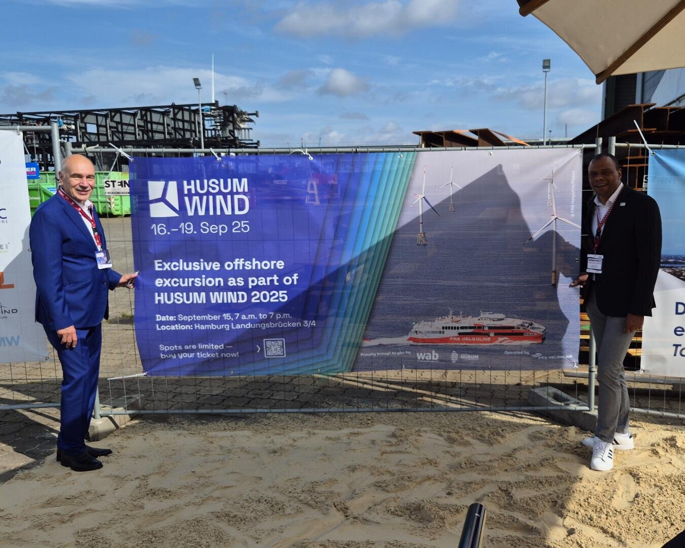 Two men in business attire stand in the sand by a fence and point to a large banner advertising the Husum Wind 2025 offshore excursion. The banner features wind turbines, a ship and event information.