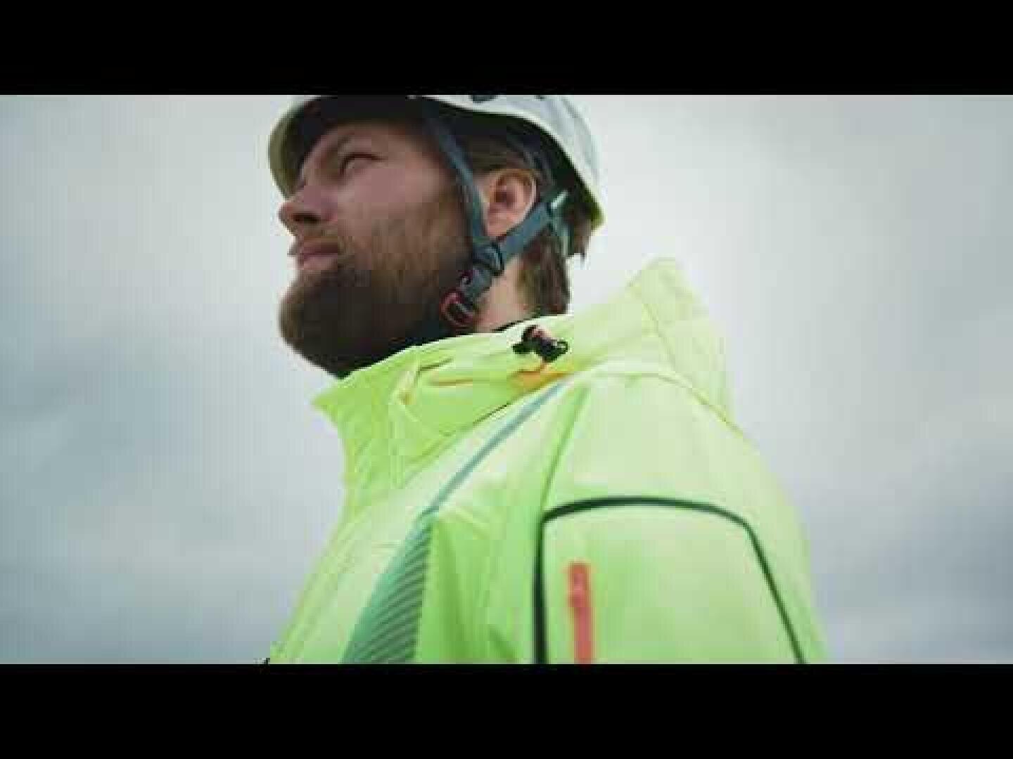 A bearded person wearing a white safety helmet and a bright yellow reflective jacket looks thoughtfully into the distance against a cloudy sky.