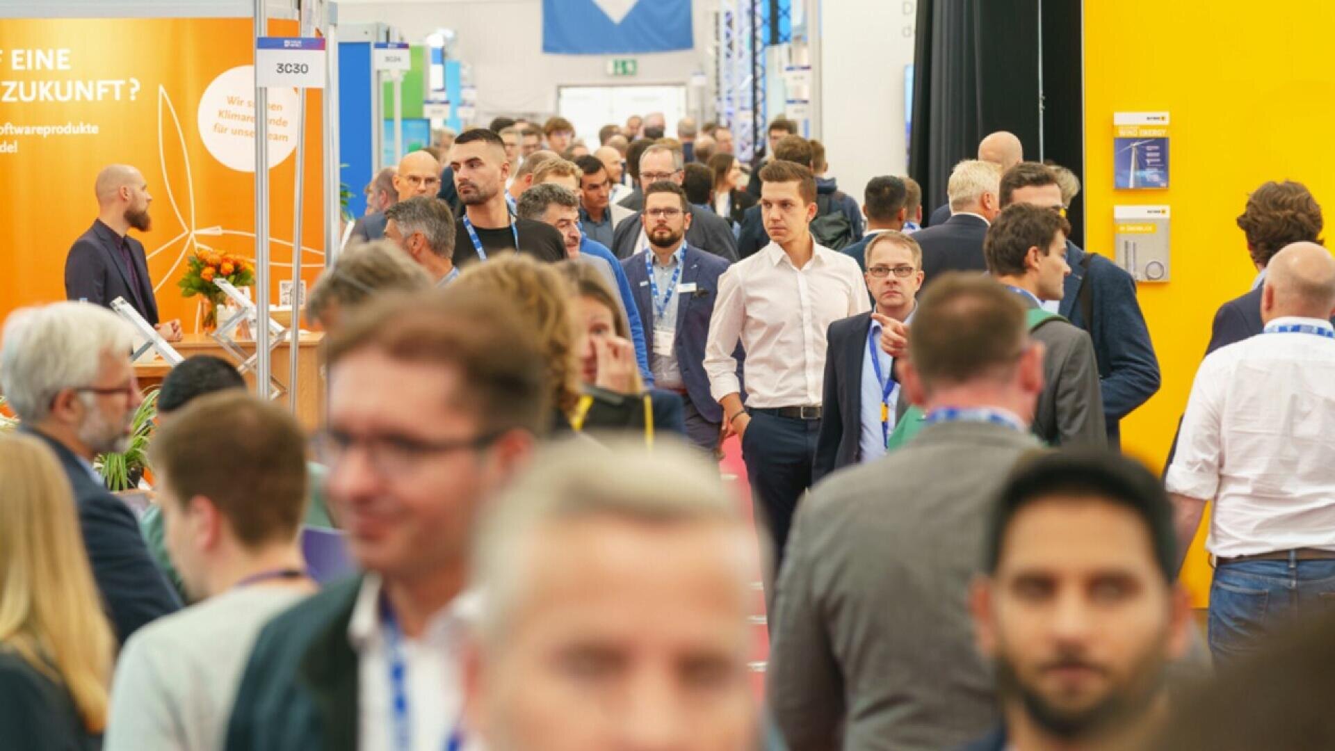 A crowded trade show or conference hallway filled with people walking and talking; colorful booths and banners line both sides, and many attendees wear conference badges around their necks.