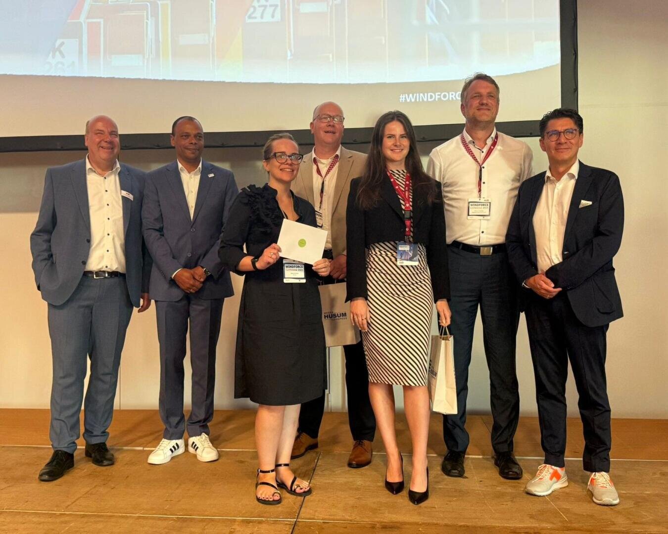 Sieben Personen in Business-Kleidung stehen auf einer Bühne und lächeln für ein Gruppenfoto. Zwei Frauen im Vordergrund halten Urkunden oder Taschen. Im Hintergrund sind eine große Leinwand und die Veranstaltungsbeschilderung zu sehen.