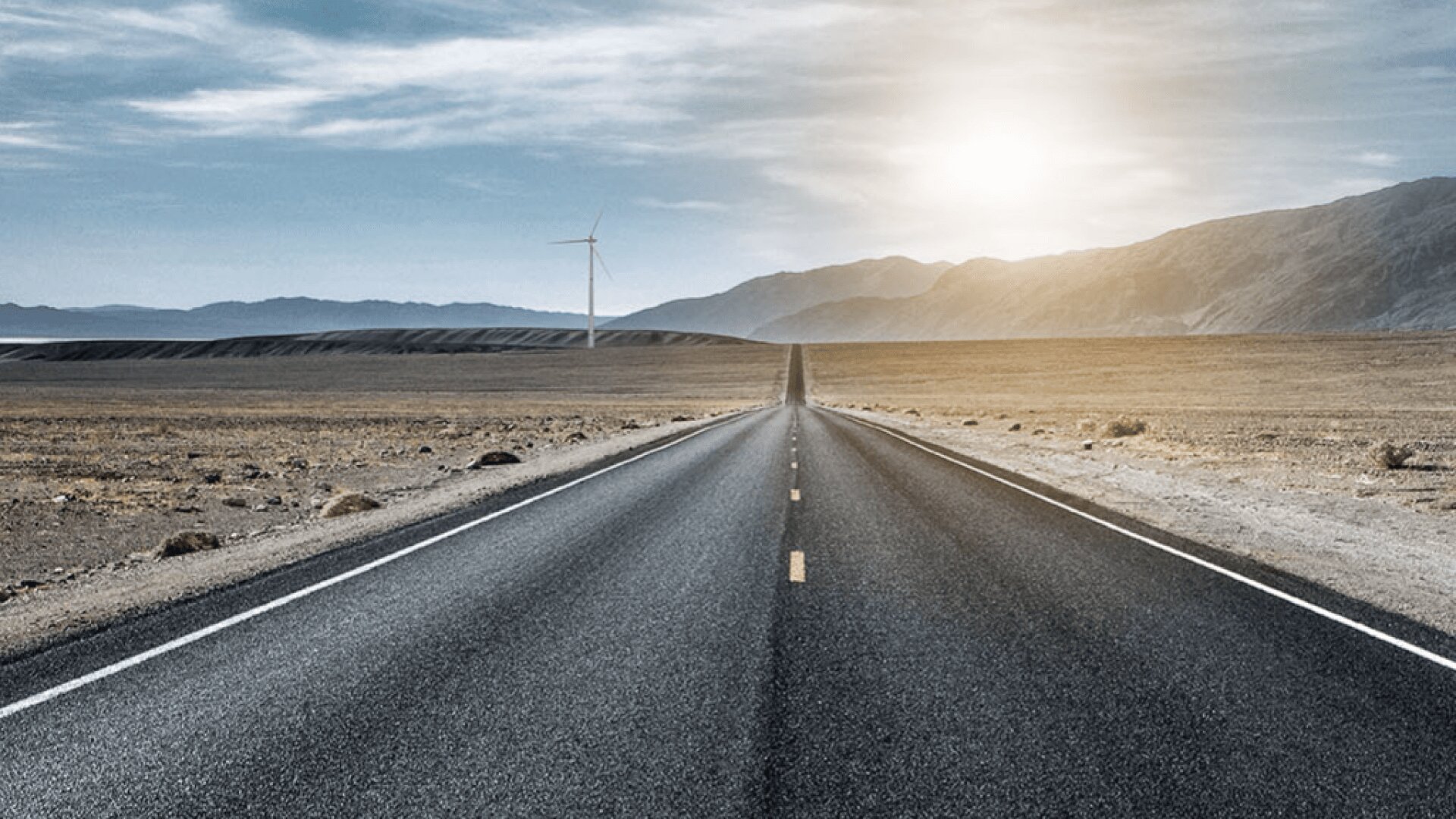 A straight, empty road stretches into the distance through a barren desert landscape with mountains on the horizon, a wind turbine to the left, and the sun shining through a partly cloudy sky.