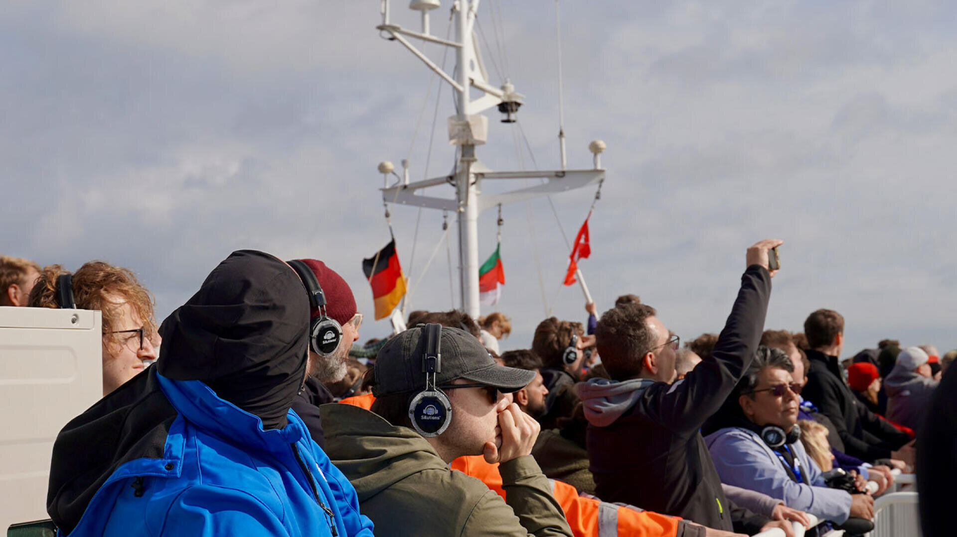 Eine Menschenmenge auf dem Bootsdeck, die Kopfhörer trägt und zum Horizont blickt.