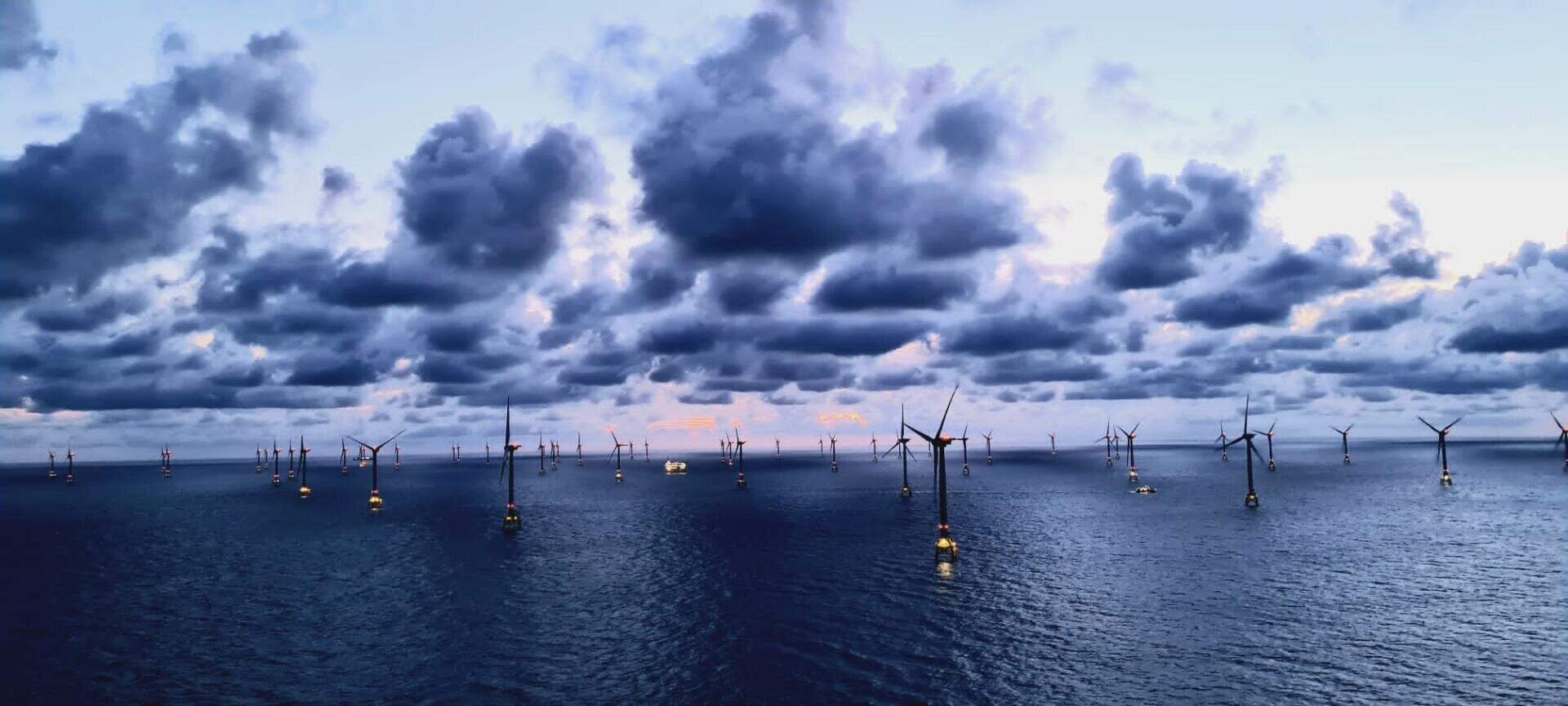 Reihen von Offshore-Windturbinen stehen verstreut auf dem ruhigen Meer unter einem dramatischen, wolkenverhangenen Himmel in der Abenddämmerung, mit leuchtenden Lichtern auf den Turbinen und einem Hauch von Rosa am Horizont.