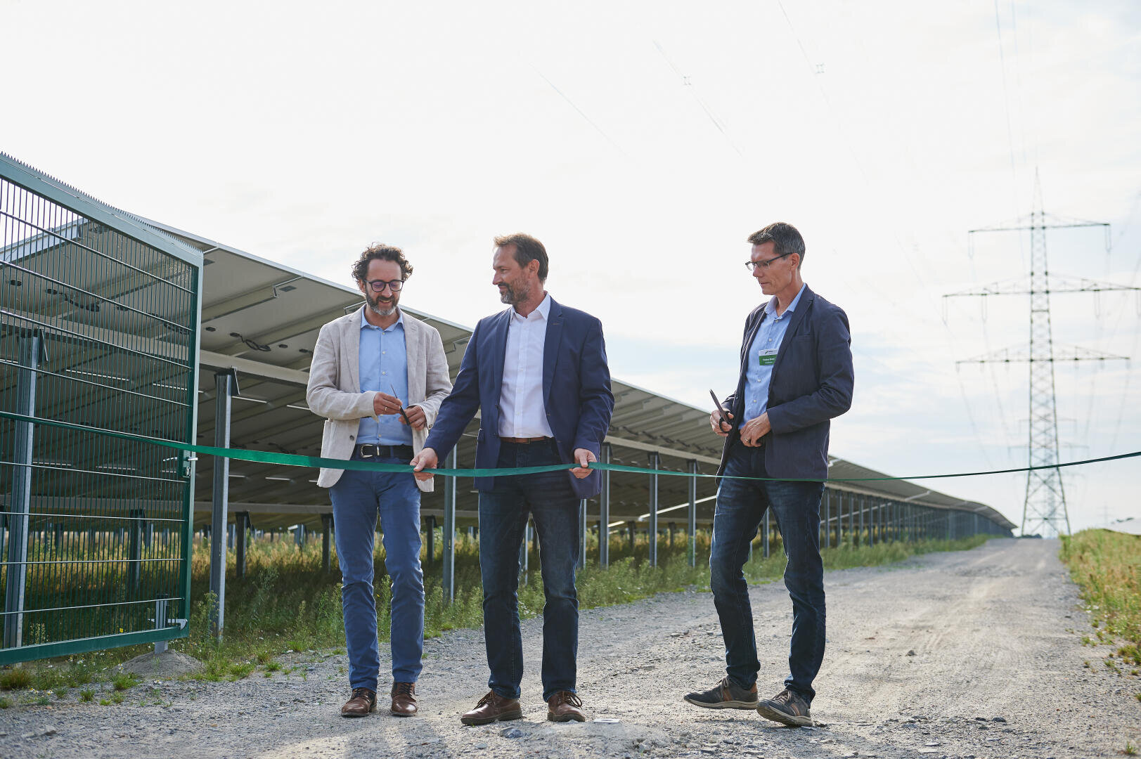 Drei Männer in Geschäftskleidung stehen auf einem unbefestigten Weg neben Solarmodulen. Einer von ihnen schneidet mit einer Schere ein grünes Band durch, wahrscheinlich bei einer Veranstaltung zum Durchschneiden des Bandes an einem sonnigen Tag im Freien.
