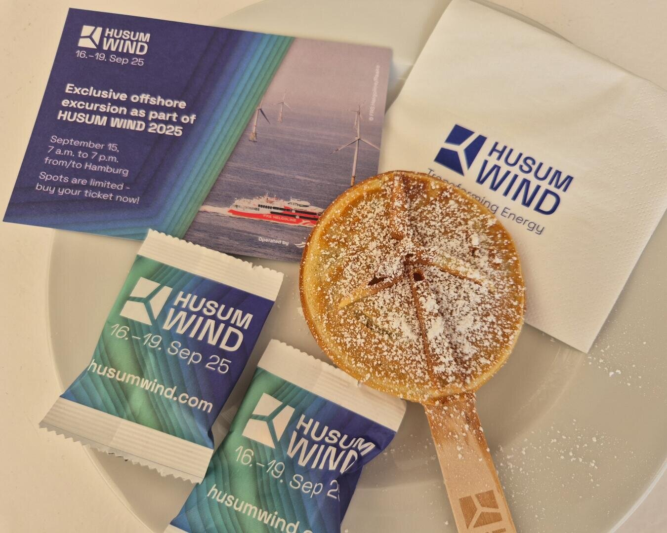 A plate with a powdered sugar cake on a wooden stick, two sweets wrapped in HUSUM WIND, a branded napkin and a flyer for an offshore excursion as part of HUSUM WIND 2025 with wind turbines and a ship.