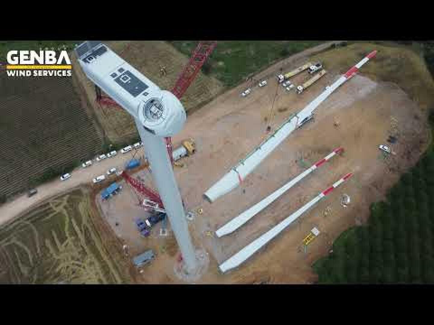 Luftaufnahme einer Windturbinen-Baustelle mit einem größtenteils montierten Turm, mehreren Turbinenflügeln am Boden, Baufahrzeugen und über eine gerodete Fläche verstreuten Geräten. Das Logo von GENBA WIND SERVICES ist in der oberen linken Ecke zu sehen.