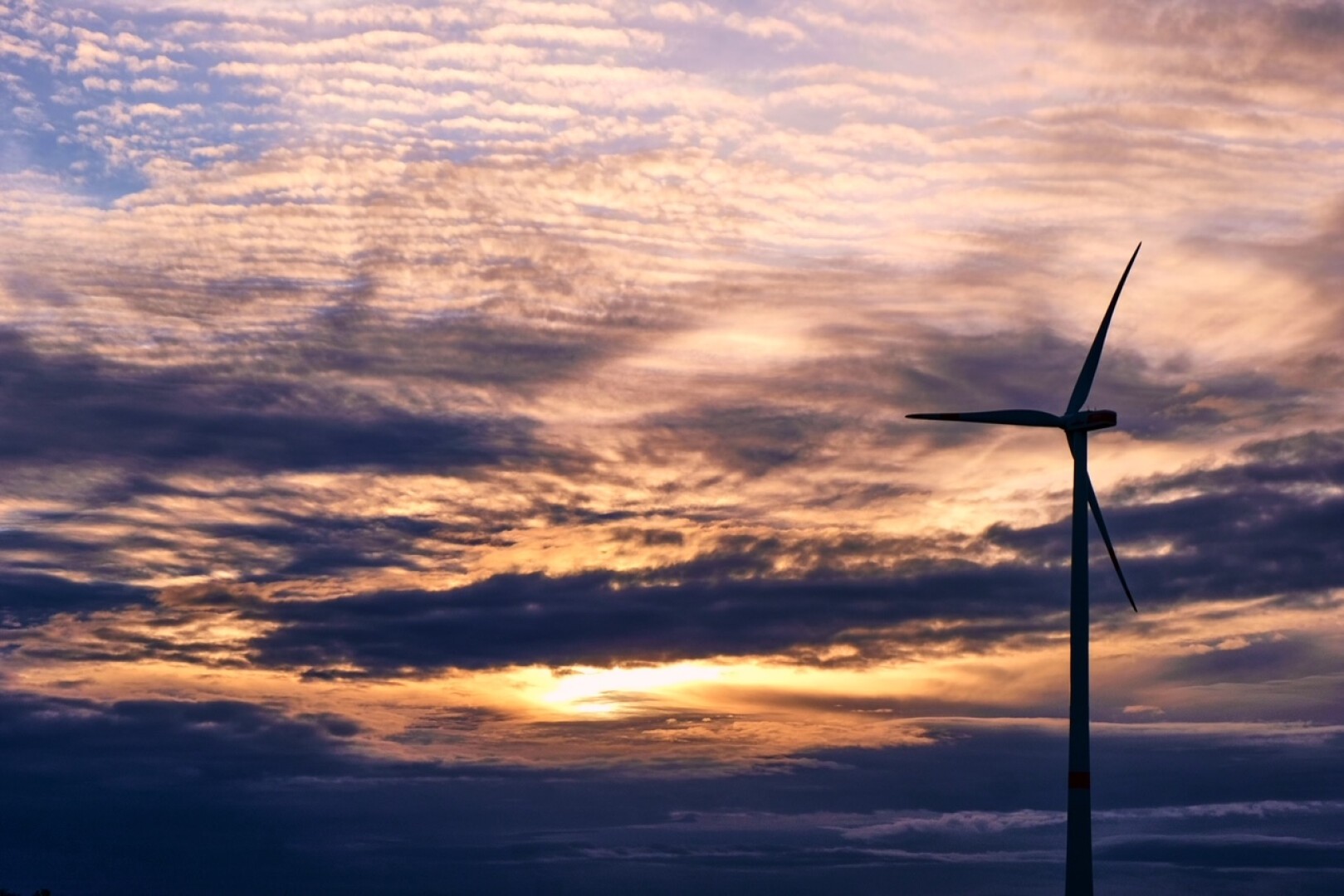 A wind turbine stands against a dramatic sunset sky filled with layers of clouds in shades of orange, yellow, purple, and blue.