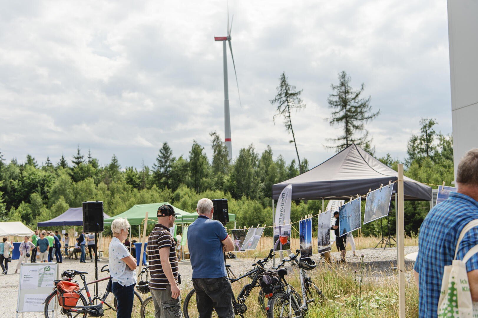 Menschen versammeln sich im Freien in der Nähe von Zelten und Schautafeln, in der Nähe sind Fahrräder geparkt. Im Hintergrund steht eine Windkraftanlage vor einem teilweise bewölkten Himmel und einem Waldgebiet.