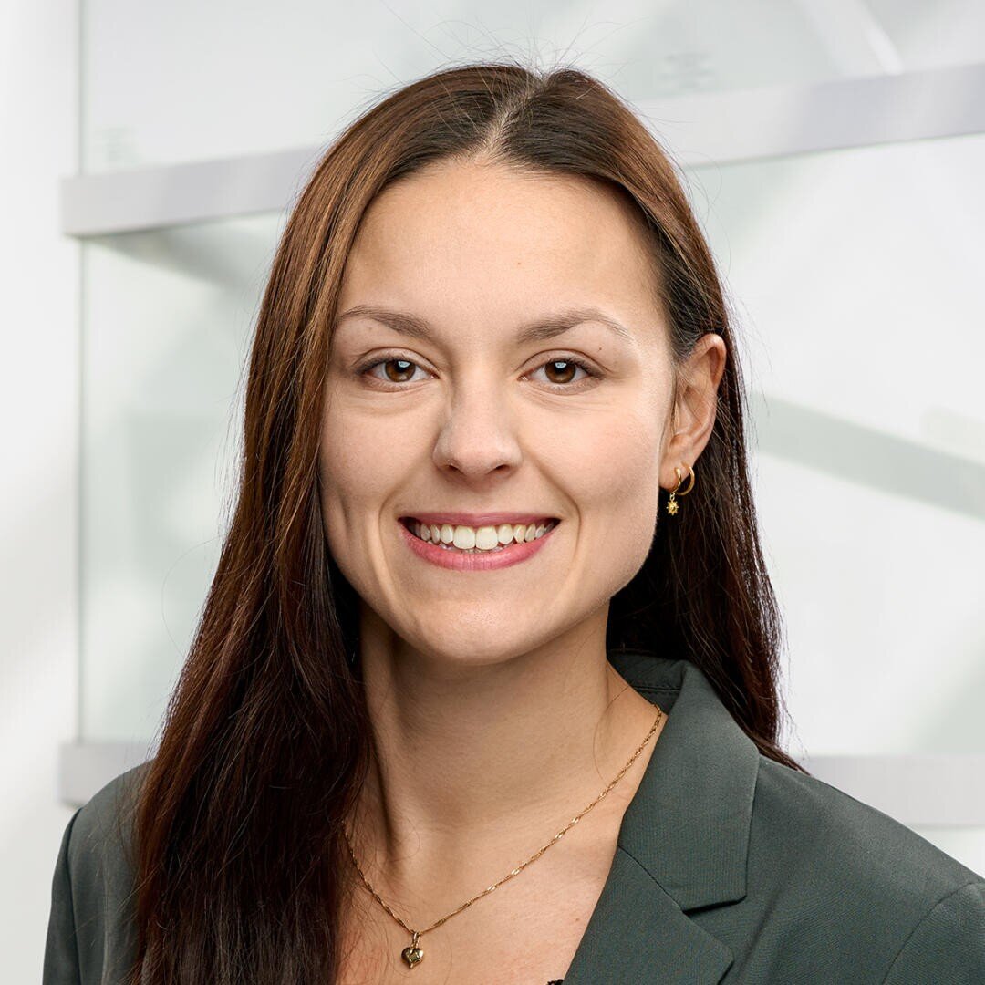 A woman with long brown hair, wearing a dark blazer, small hoop earrings, and a gold necklace, smiles at the camera against a light, modern background.