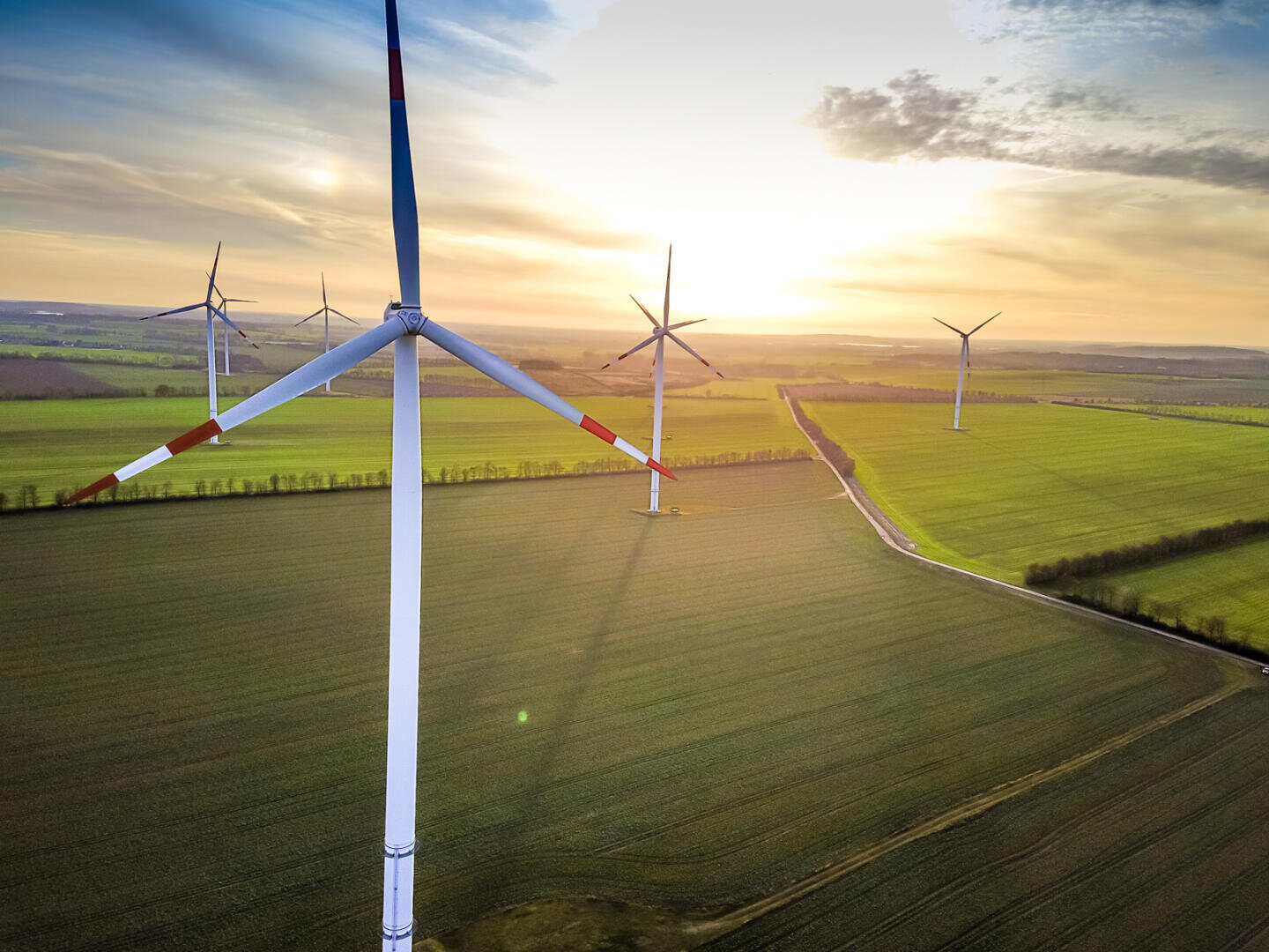 Luftaufnahme von Windturbinen auf weitläufigen grünen Feldern bei Sonnenuntergang, wenn der Himmel teilweise bewölkt ist und sanftes Sonnenlicht die Landschaft beleuchtet.