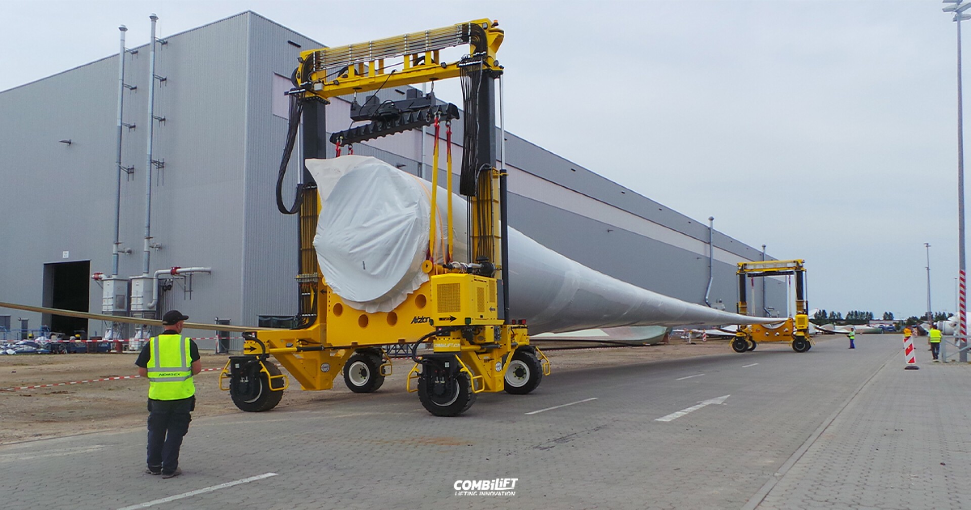 Zwei gelbe Transportmaschinen transportieren ein großes Windradblatt horizontal vor einem Industriegebäude. Arbeiter in Sicherheitswesten stehen daneben und steuern die Maschine.