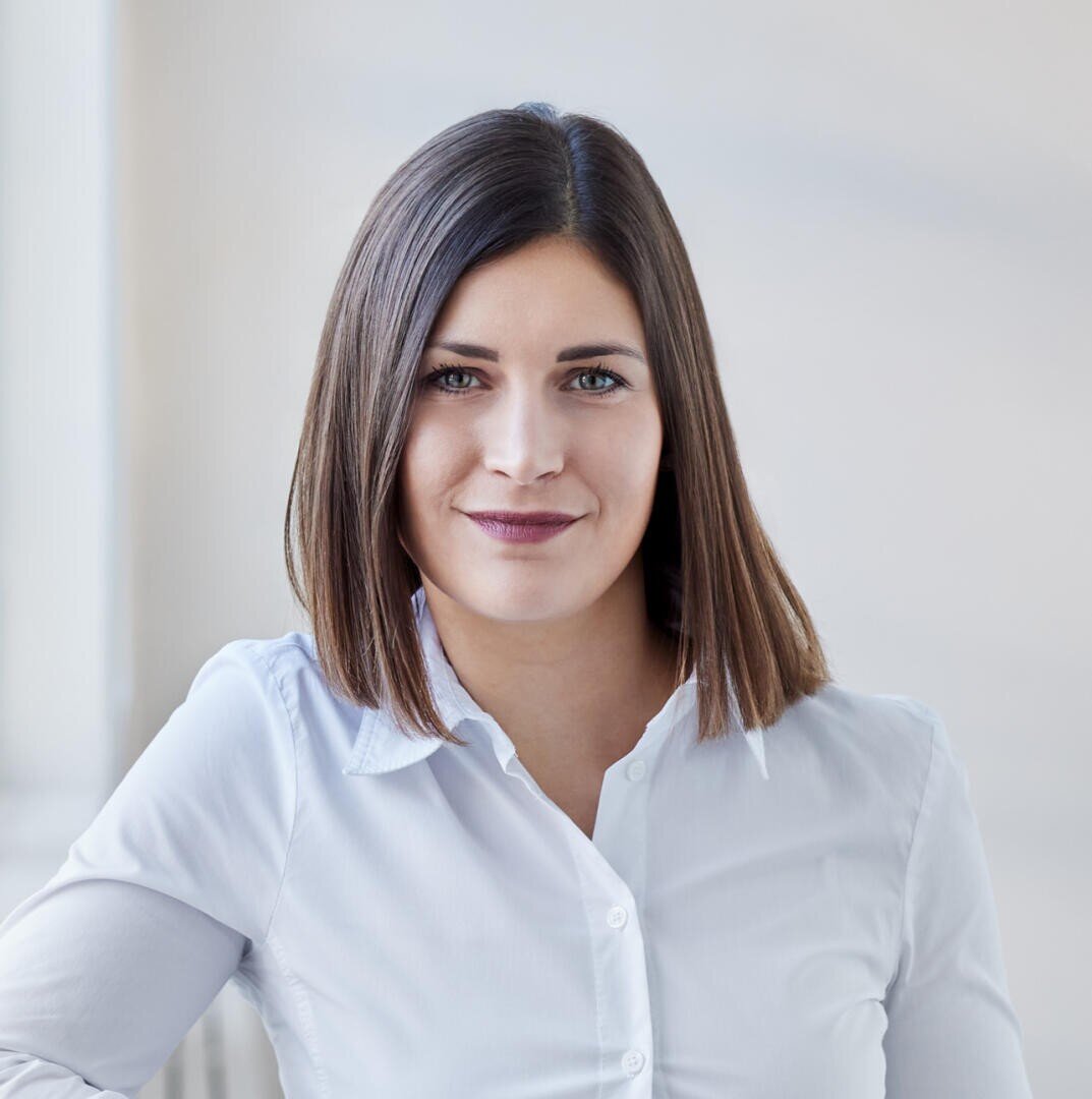 A woman with straight, shoulder-length brown hair is smiling slightly. She is wearing a white button-up shirt and stands against a blurred light background.
