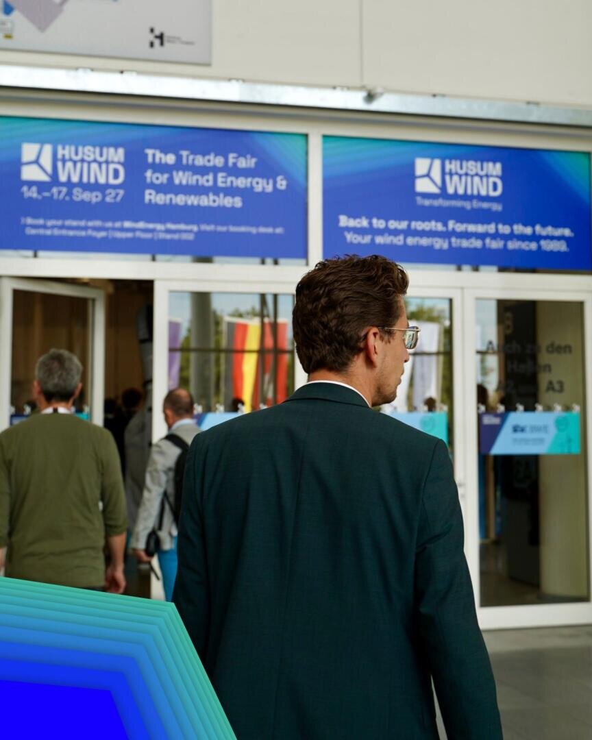 Ein Mann im Anzug steht am Eingang einer Messe für Windenergie und erneuerbare Energien; über den Türen hängen blaue Banner, und mehrere Menschen betreten das Messegelände.