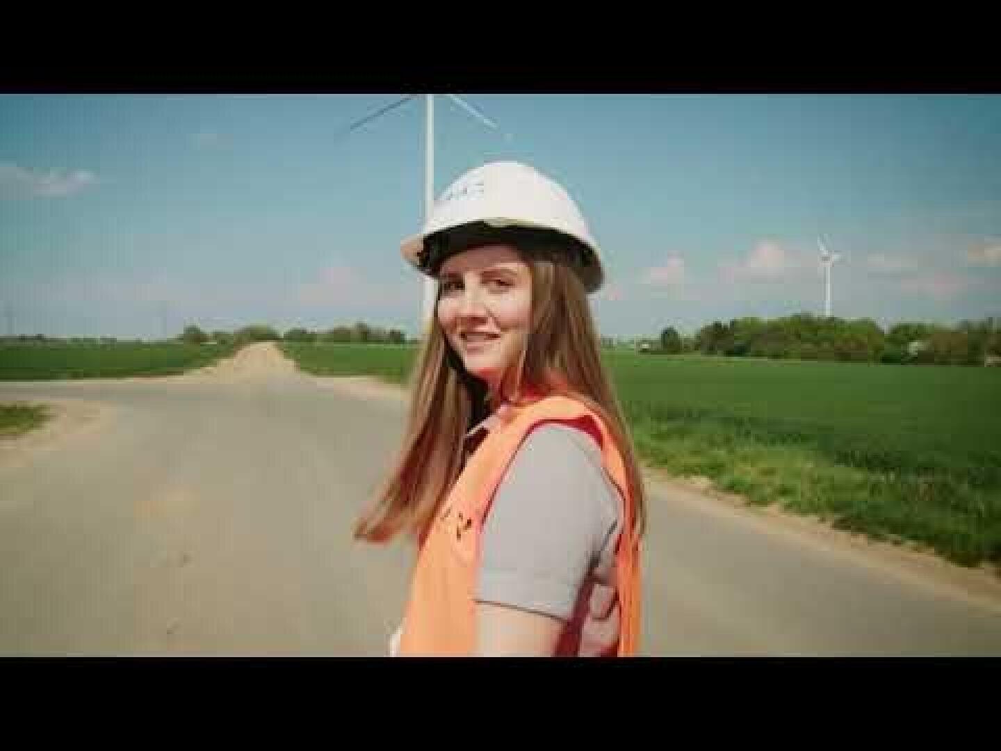 Eine junge Frau mit einer orangefarbenen Sicherheitsweste und einem weißen Schutzhelm steht auf einer Landstraße und lächelt in die Kamera. Im Hintergrund sind grüne Felder und Windkraftanlagen unter blauem Himmel zu sehen.
