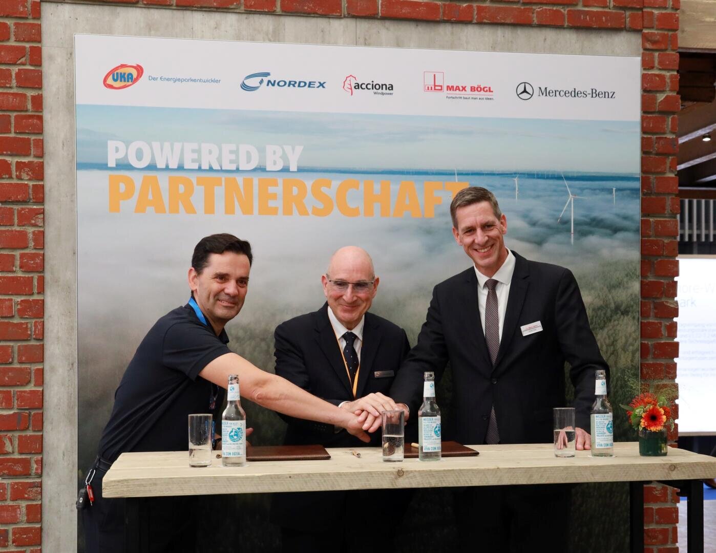 Drei Männer in Geschäftskleidung stehen hinter einem Tisch, schütteln Hände und lächeln für ein Foto. Hinter ihnen ist ein Banner mit der Aufschrift "Powered by Partnerschaft" und Logos verschiedener Unternehmen zu sehen. Auf dem Tisch stehen Wasserflaschen und eine Blumenvase.