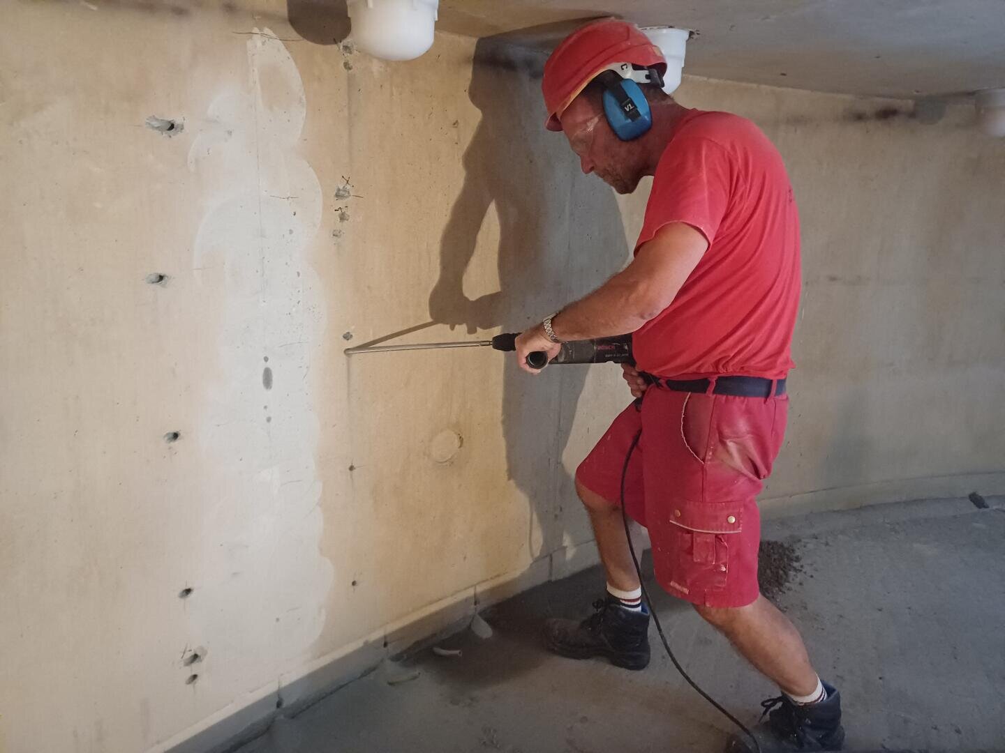 Ein Bauarbeiter in roter Kleidung, mit Schutzhelm und Gehörschutz arbeitet mit einer Bohrmaschine an einer Betonwand im Inneren eines Gebäudes.