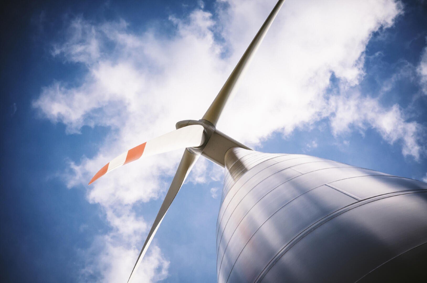 Eine Nahaufnahme einer Windturbine vor einem blauen Himmel mit vereinzelten weißen Wolken, die die glatte Metallstruktur und die Flügel zeigt, von denen einer mit roten Streifen versehen ist.