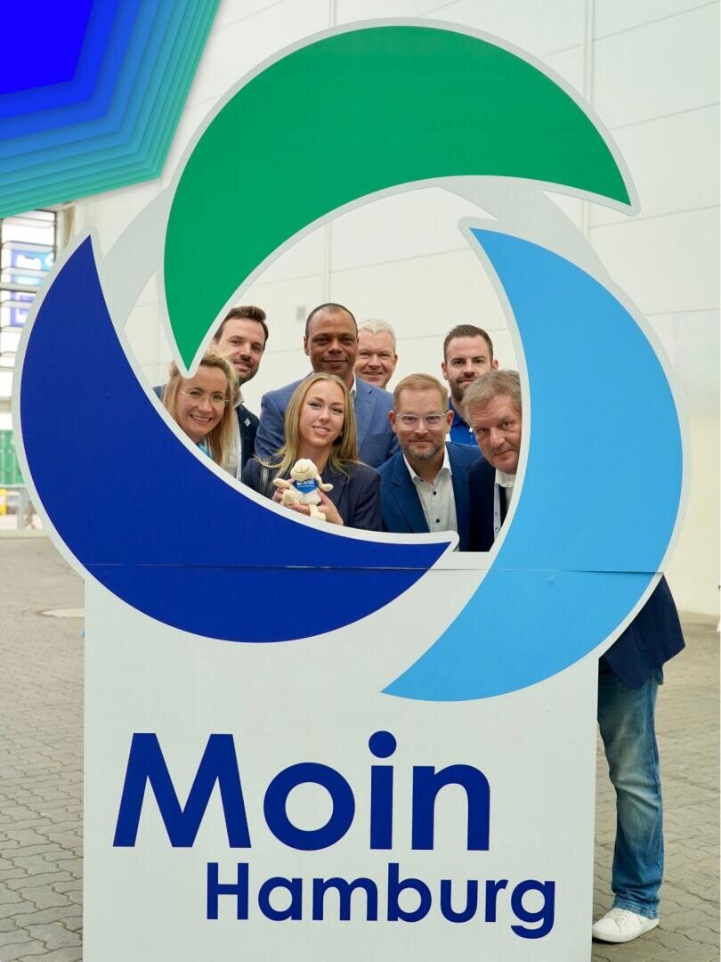 Acht Personen posieren hinter einem großen Schild mit blauen und grünen Kreisen und der Aufschrift „Moin Hamburg“ in fetten Buchstaben in einem hellen, modernen Gebäude.