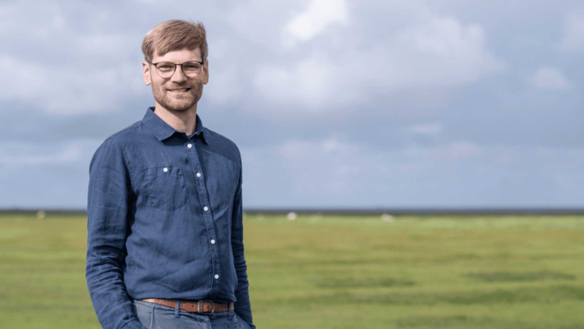 A man with glasses and a beard, wearing a blue button-up shirt and gray trousers, stands smiling in an open meadow under a cloudy sky.