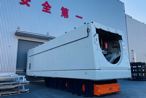 Ein großer, weißer Waggon steht auf einem orangefarbenen Bahnsteig vor einem grauen Industriegebäude mit roten japanischen Schriftzeichen über den Türen.