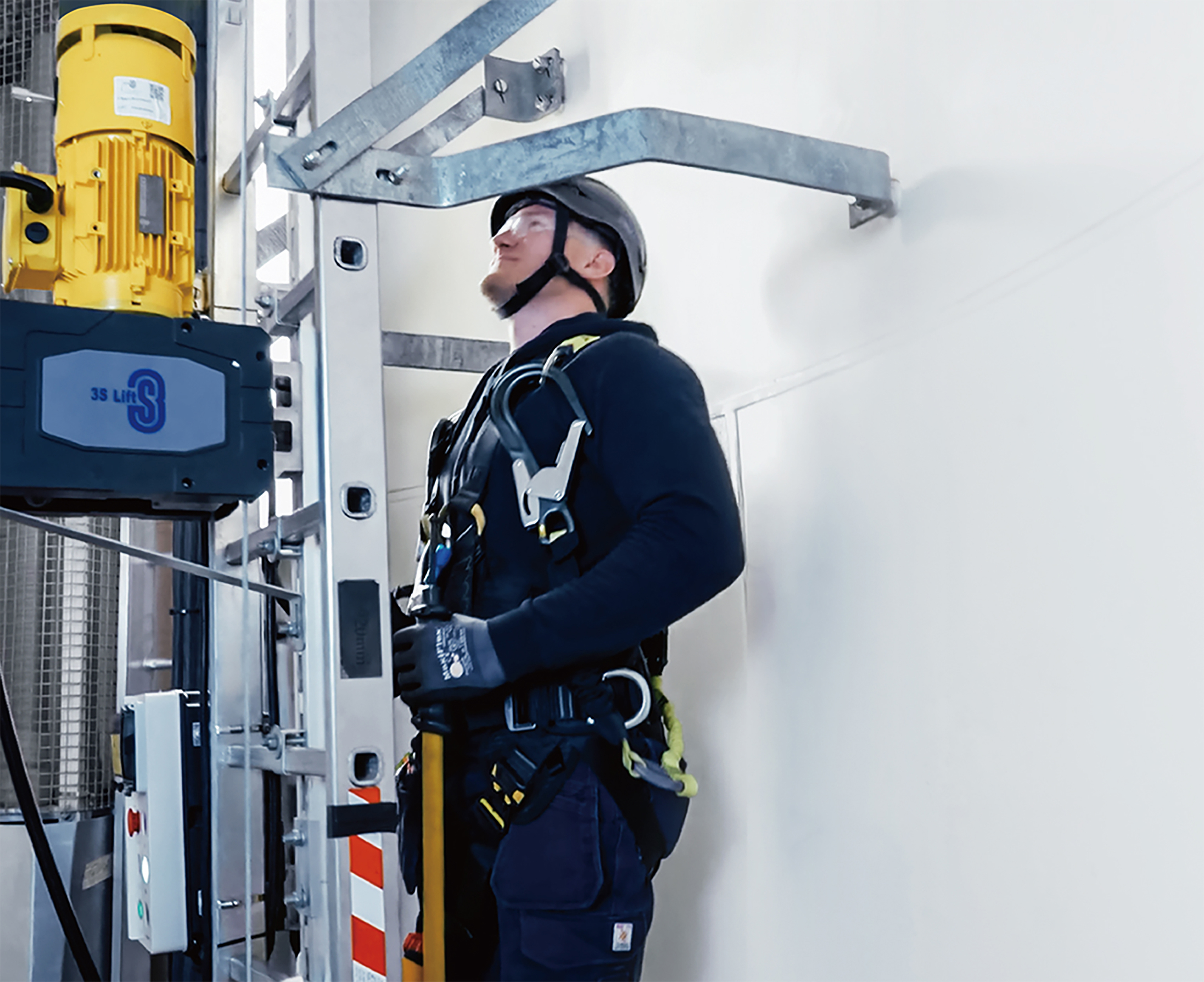 A worker wearing safety gear and a helmet stands on a ladder next to industrial equipment, secured with a harness and looking upward, inside a facility with a white wall.