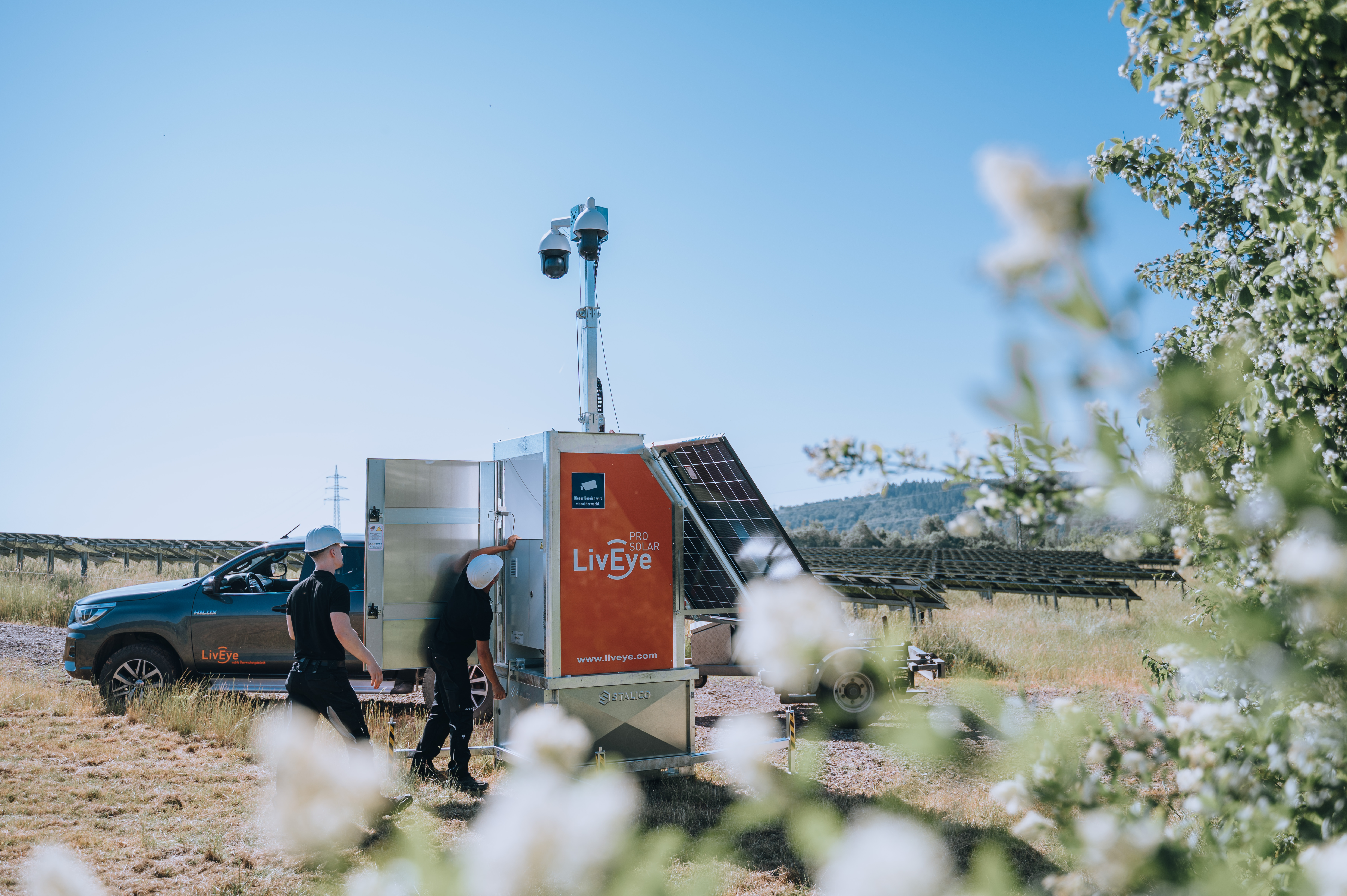 Zwei Personen mit Schutzhelmen untersuchen eine mobile Überwachungseinheit mit Solarmodulen mit der Aufschrift „Live Eye“ auf einer Wiese in der Nähe eines schwarzen Pickups, im Hintergrund sind Stromleitungen und Solarmodule zu sehen.