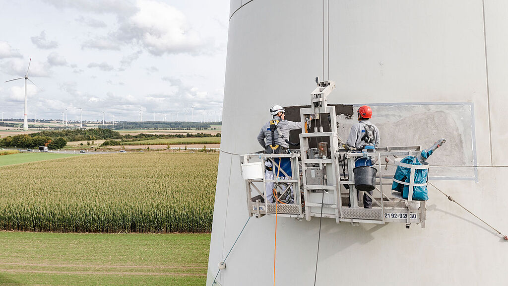 Zwei Arbeiter in Schutzkleidung befinden sich auf einer aufgehängten Plattform und inspizieren oder reparieren die Seite einer großen Windkraftanlage. Im Hintergrund ist eine ländliche Landschaft mit Feldern und weiteren Windkraftanlagen zu sehen.