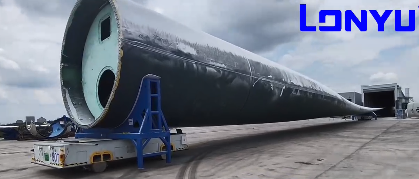 Ein großer Windturbinenflügel wird auf einem Pritschenwagen über ein Industriegelände transportiert. Der Himmel ist bewölkt, und das Wort Lonyu ist in blauer Schrift in der oberen rechten Ecke zu sehen.