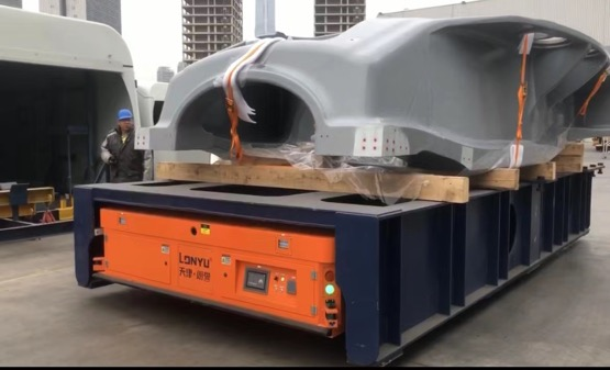 Ein großes, graues Industrieteil ist auf einem blau-orangefarbenen fahrerlosen Transportfahrzeug (FTS) befestigt, auf dem die Marke LONYU zu sehen ist. Ein Arbeiter mit einem blauen Helm steht in der Nähe, und im Hintergrund sind Hochhäuser zu sehen.
