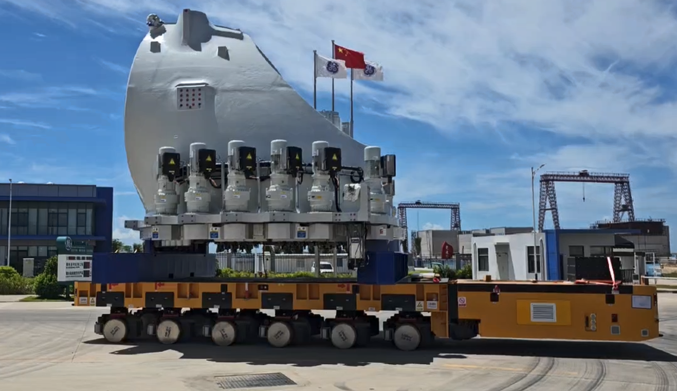 A large, heavy industrial component is transported on a multi-wheeled yellow vehicle at an industrial site under a blue sky, with Chinese flags and buildings visible in the background.