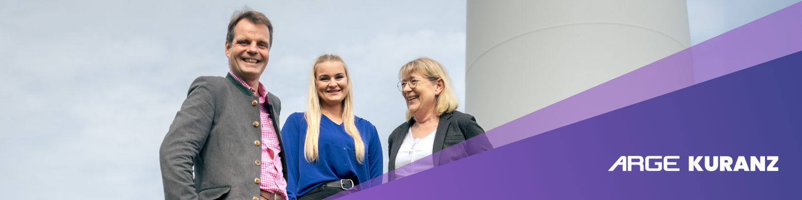 Drei Personen, zwei Frauen und ein Mann, stehen lächelnd im Freien vor einer weißen Struktur und einem teilweise bewölkten Himmel. Das Bild ist mit einem violetten Overlay mit dem Text ARGE KURANZ in der unteren rechten Ecke versehen.