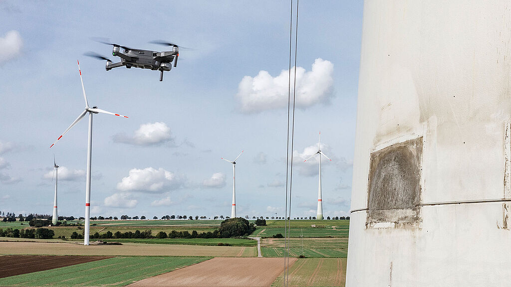 Eine Drohne fliegt in der Nähe einer großen Windkraftanlage in einer ländlichen Landschaft mit Feldern und mehreren anderen Windkraftanlagen unter einem teilweise bewölkten Himmel.